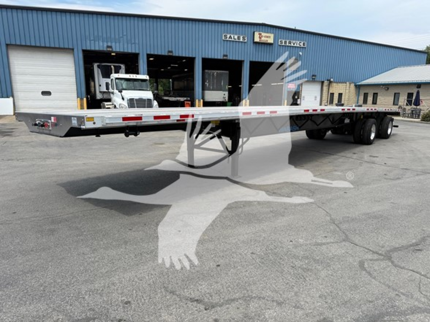 Flatbed trailer showing long aluminum deck with red/white reflective rails and tandem rear axles in front of a blue sales building.