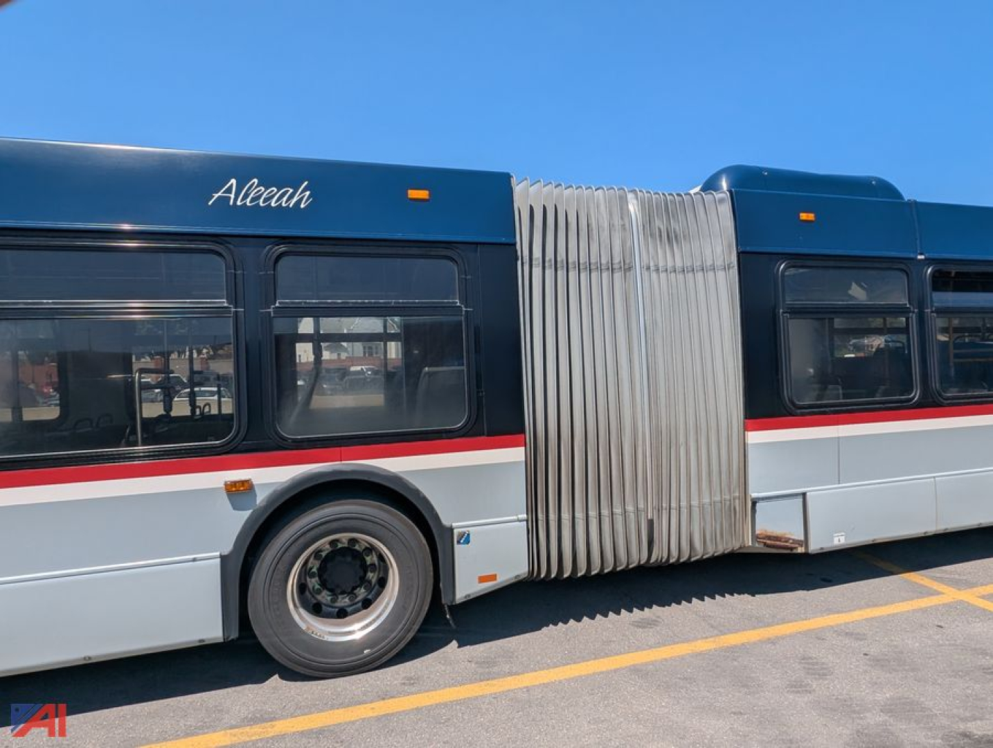 Transit bus side view showing accordion accordion-style bus connection with folding hallway. Visible text 'Aleah' on blue upper panel, grey side with red stripe, wheel and tire, mid-section bellows accordion joint.