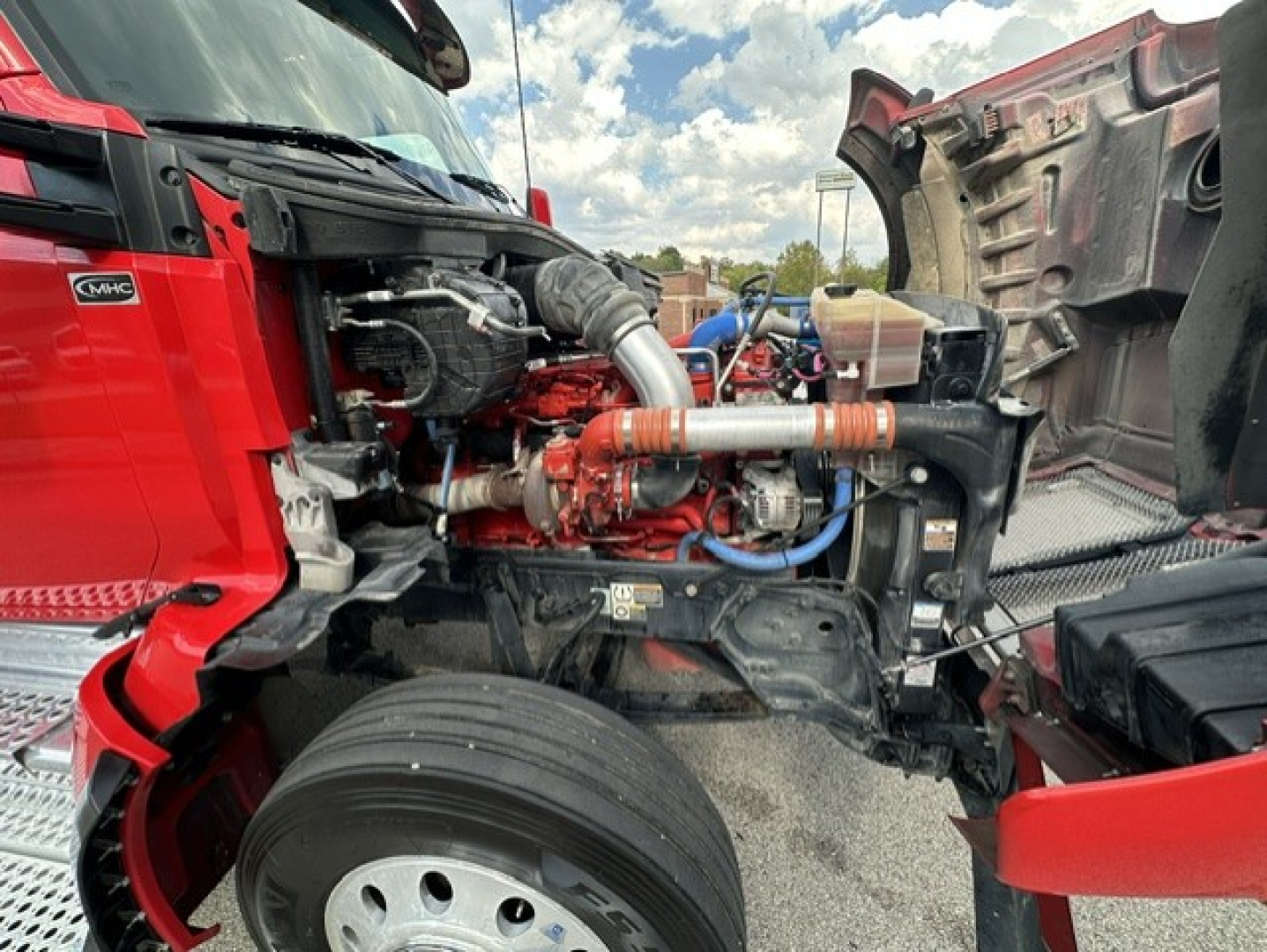 Kenworth T880 engine bay with exposed Cummins engine components, orange intake piping, blue hoses, and red truck frame visible on left; hood open exposing engine and front wheel.