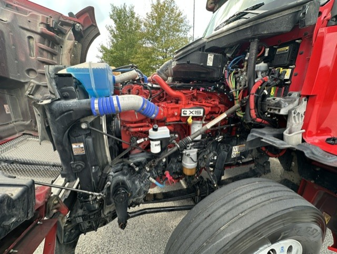 Kenworth T880 engine bay showing Cummins X15 engine with red blocks, blue silicone hose, various fluid reservoirs, and front wheel visible.