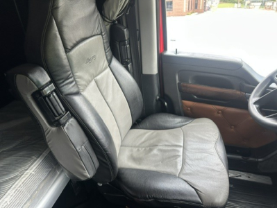 Truck cab interior seat and door panel, black leather seat with embossed 'KENWORTH' logo, tan door panel trim, steering wheel visible.