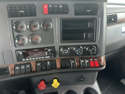 Dashboard of a Kenworth T880 cab showing multiple gauges, switch panels, and climate controls. Prominent red hazard button, square cabin air vents, and woodgrain trim.