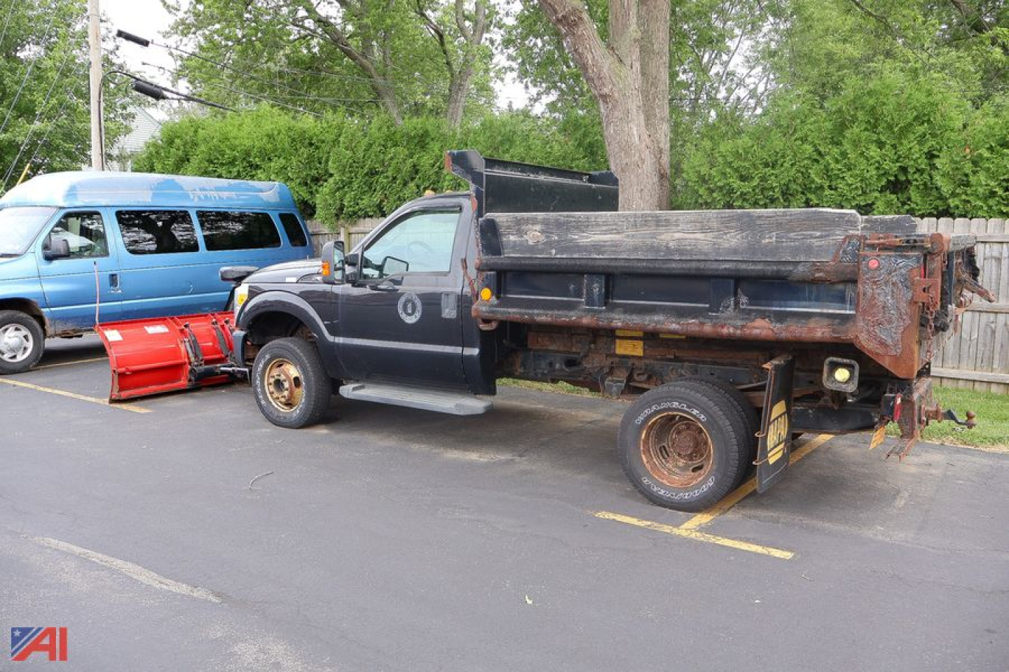 Ford F-350 Super Duty dump bed truck with plow attachment in a parking lot. Dirt on bed, dual rear wheels, rusted rims, and a detached red snow plow blade on the ground nearby.