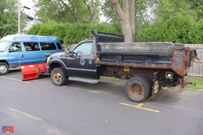 Ford F-350 Super Duty dump bed truck with plow attachment in a parking lot. Dirt on bed, dual rear wheels, rusted rims, and a detached red snow plow blade on the ground nearby.