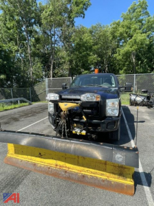 Front view of a black Ford F-350 Super Duty pickup with a snow plow and lift gate attached in a parking lot; yellow plow blade in the foreground, orange beacon light on roof, trees and chain-link fence in background.