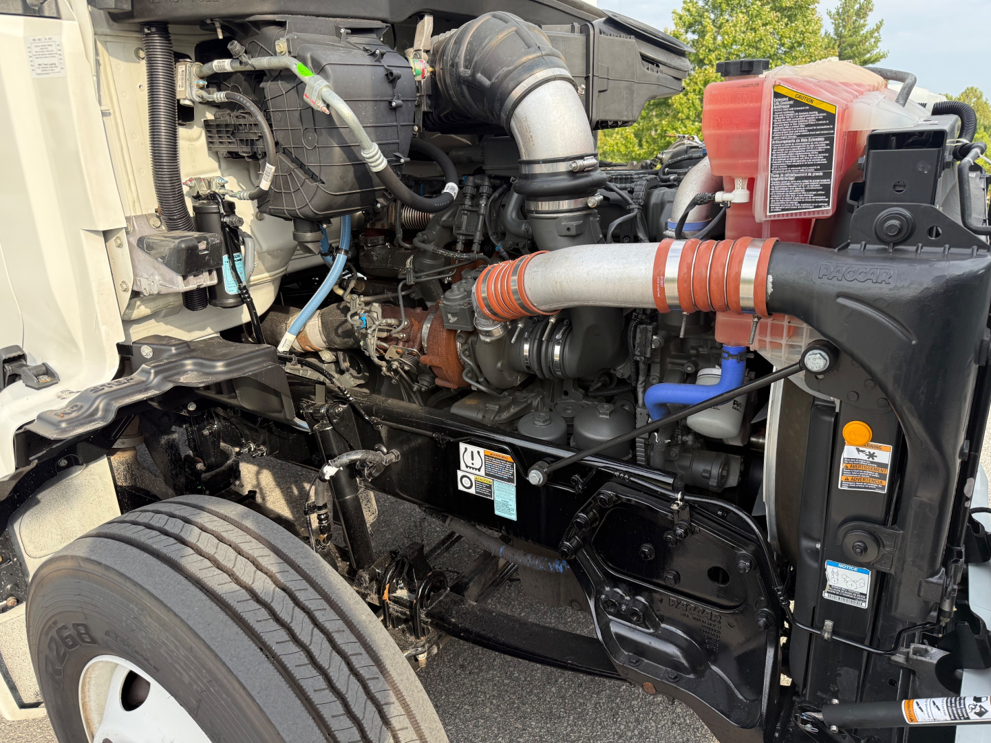 Kenworth T680 engine bay showing air intake, coolant reservoir, hoses, and battery area. Visible components include intake tubing, turbo piping, and front wheel well.