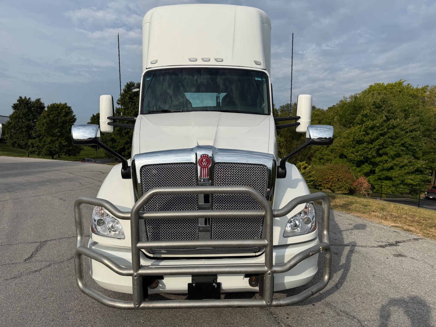 Front view of a white Kenworth T680 with a heavy-duty grille guard. Large chrome grille, emblem in center, dual rectangular headlights, side mirrors, and aerodynamic hood.
