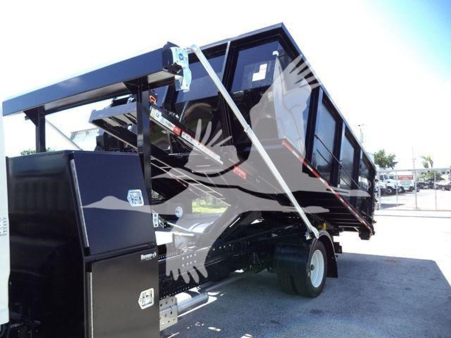 Isuzu roll-off truck body with black dump bed raised, side panels, hydraulic link, and wheel. Some text visible on the tank and decals but not fully legible.