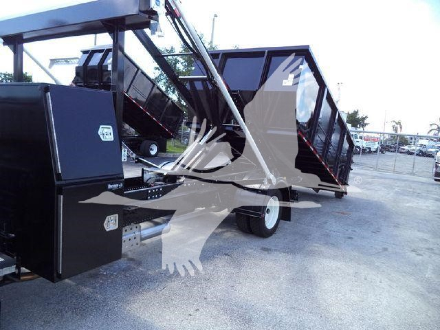 Truck body in-rolloff setup showing hydraulic lifting mechanism and cargo bed with black metal panels; mounting frame and wheels visible.