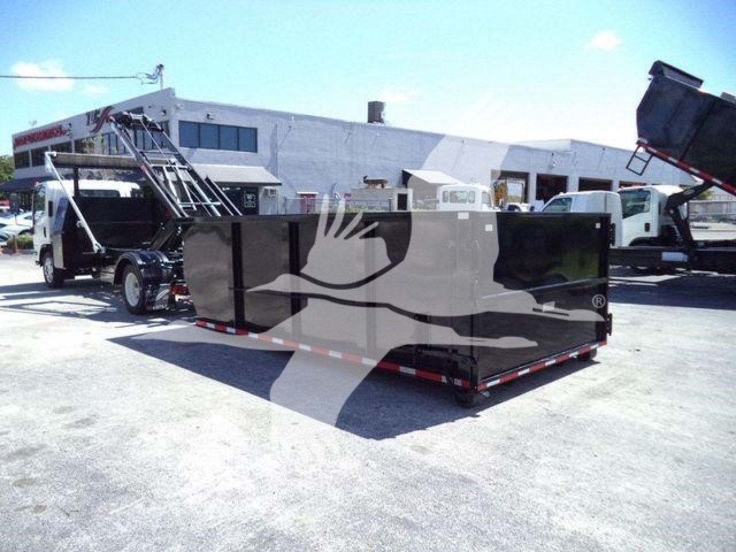 Isuzu NPRR rolloff truck in a dealership lot with a large black roll-off container loaded in bed.