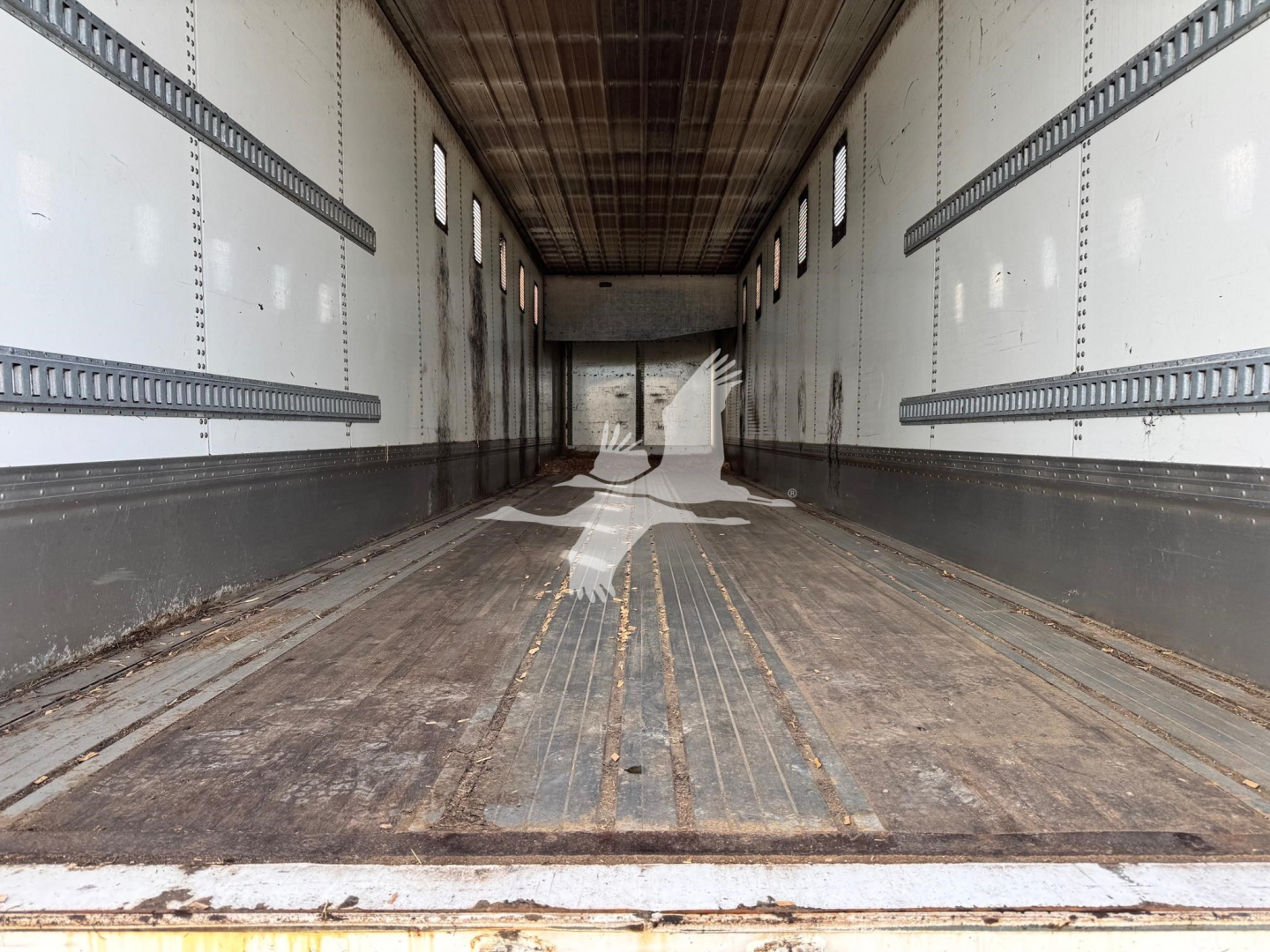 Trailer interior showing empty cargo area with ribbed steel floor and sidewalls; windows along the top; worn wooden floor accents and wheel wells at rear.