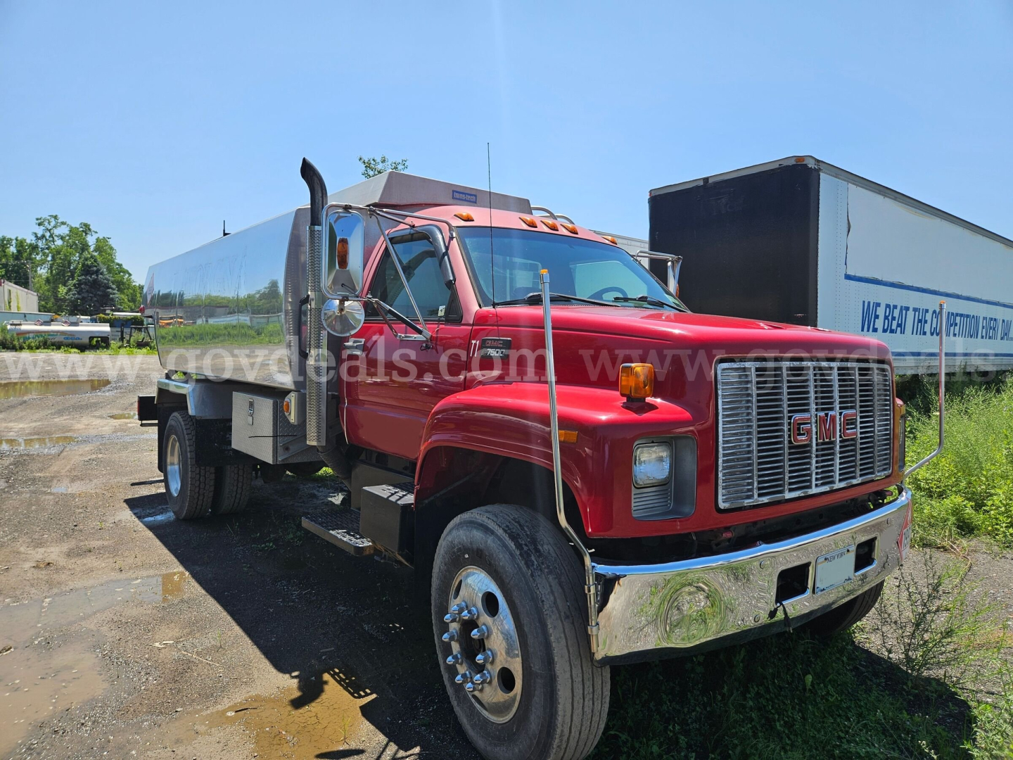 GMC C7500 truck with red cab and large stainless steel tank in side view showing GMC grille and dual rear wheels.