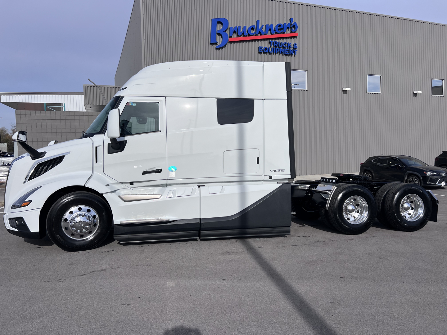 Side view of a new 2026 Volvo VNL64T840 tractor with a white cab and a tandem rear axle, parked in front of a trucking equipment building.