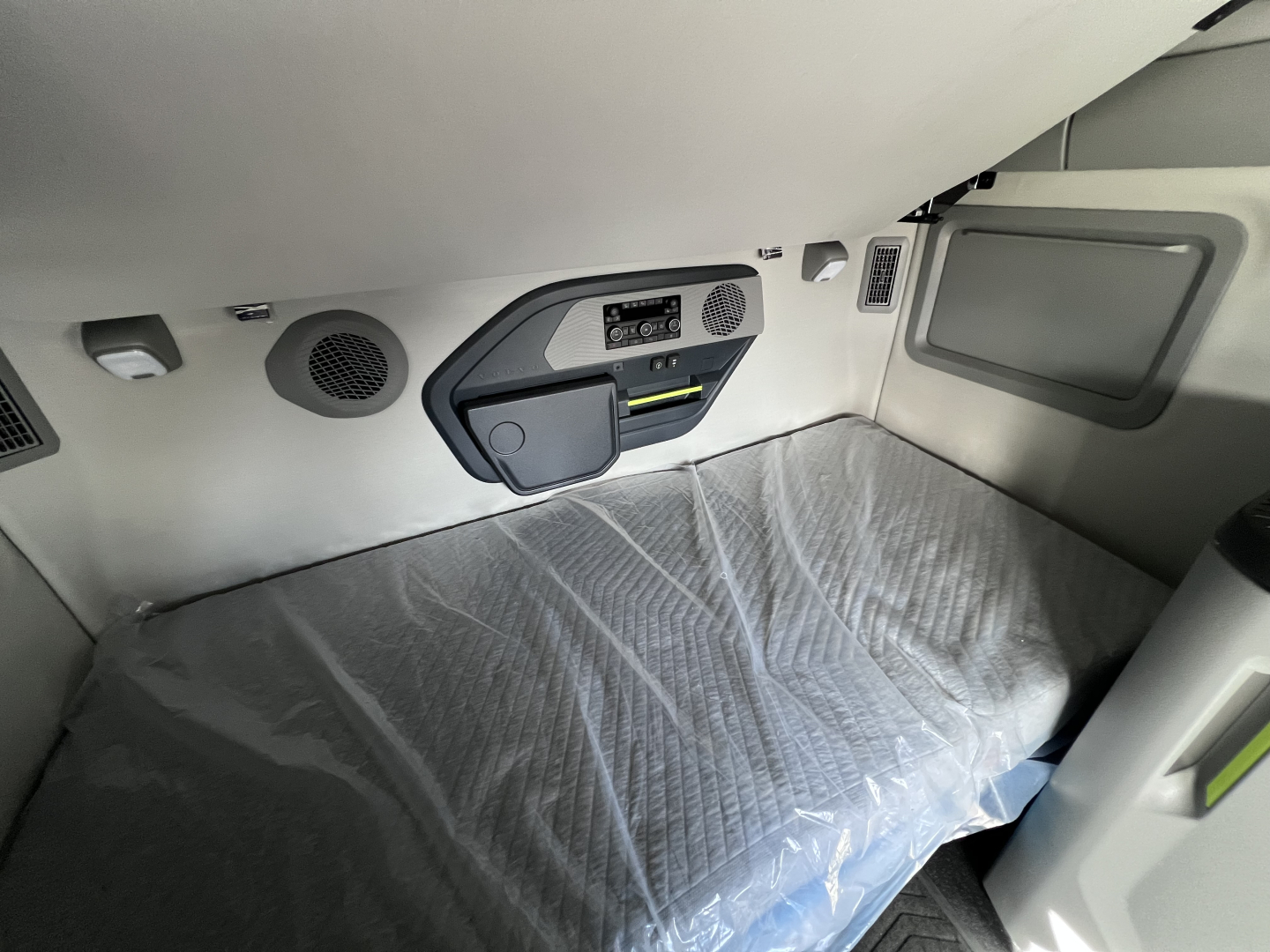 Interior sleeper cab area of a 2026 Volvo VNL64T840 showing storage panel, side window, and a covered bunk bed with protective sheet.