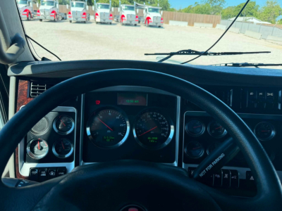 Kenworth truck cockpit dashboard view with steering wheel and gauge cluster showing speedometer and tachometer, windshield and wipers, truck lot in background.