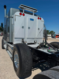 Kenworth truck rear cab area showing aluminum diamond plate compartments, rear lights, and chassis with single rear wheels. Visible text: no readable labels or serials. 