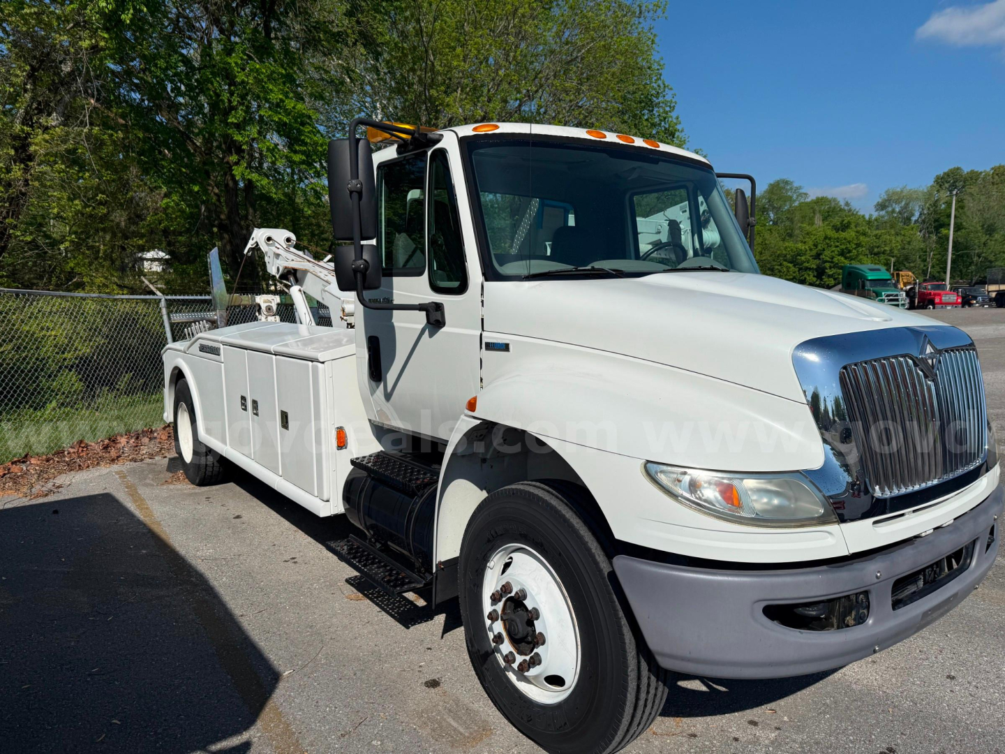 Tow truck with hook lift on International 4400, white cab, front grille, mirrors, and equipment compartments visible.