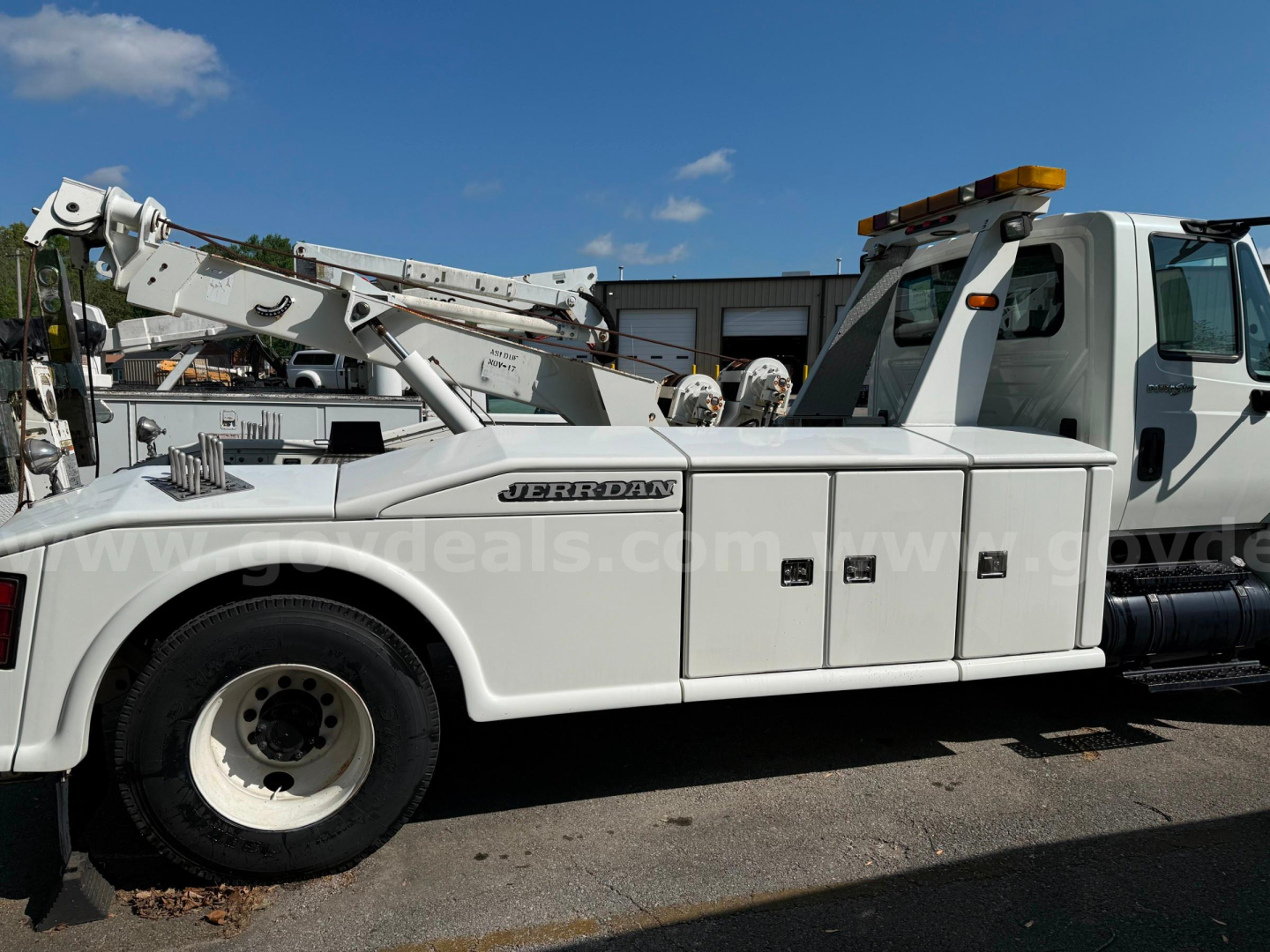 International 4400 tow truck with Jerr-Dan crane, side compartments labeled with multiple access panels, low-profile white utility body, cab with orange beacon on top, wheel and tire visible.