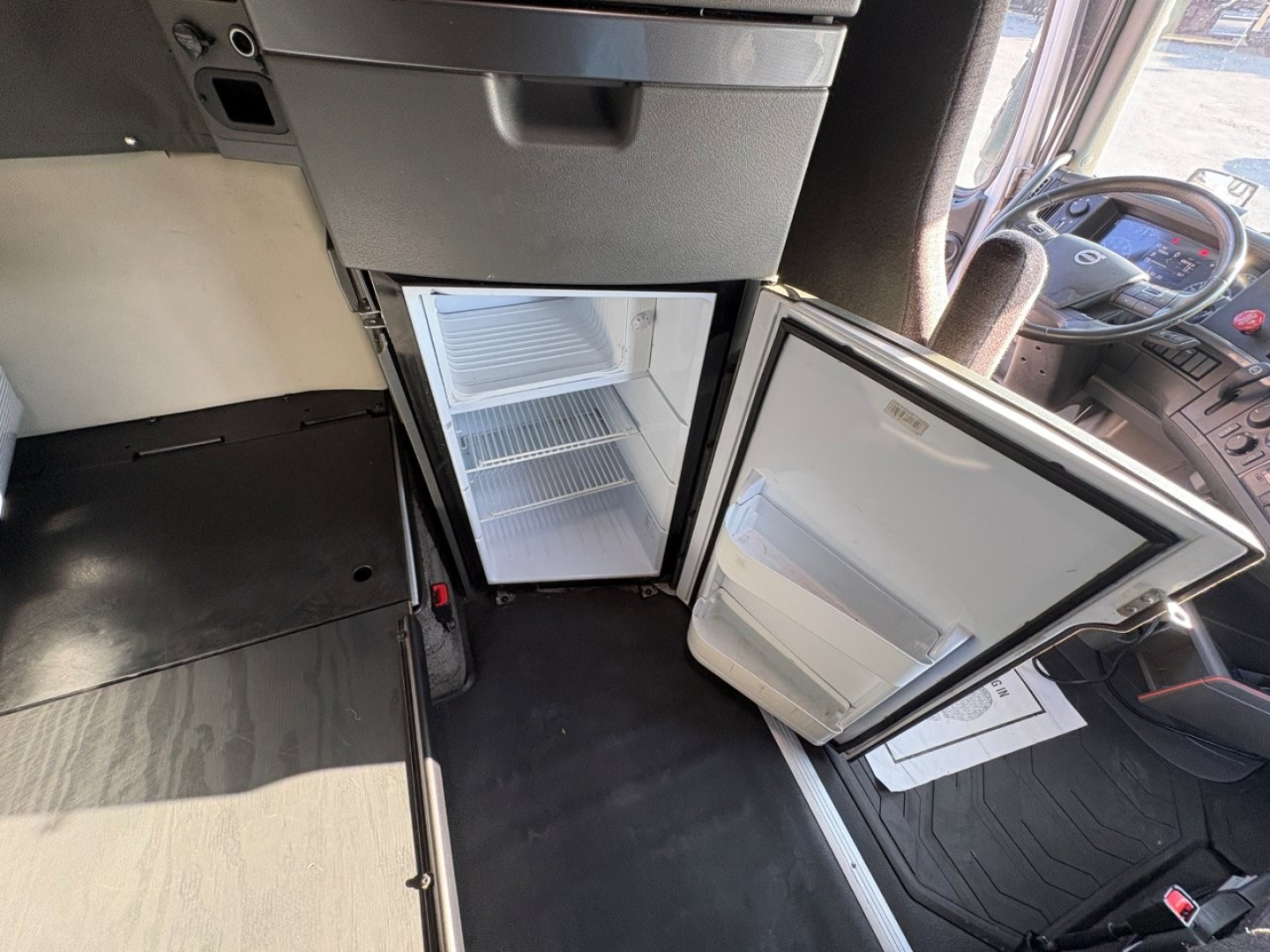 Volvo VNL64T860 cab interior showing open under-dash fridge/freezer unit and floor area with driver seat in view.