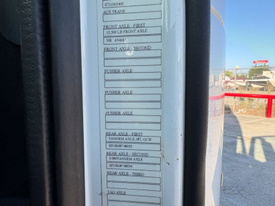 Label with multiple VIN/axle specifications on a Volvo truck door frame. Visible text includes: AUX TRANS, FRONT AXLE - FIRST 12,500 LB FRONT AXLE, NR 65463, FRONT AXLE - SECOND, PUSHER AXLE, REAR AXLE - FIRST TANDEN AXLE, 8.6CW, REAR AXLE 