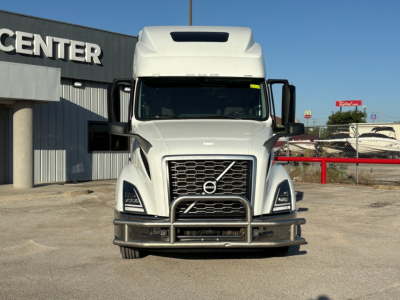 Front view of 2024 Volvo VNL64T860 truck showing grille with Volvo emblem, bumper guard, headlights, and large hood; dealership building in background.