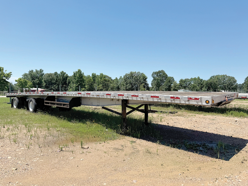 Doonan aluminum flatbed trailer in field showing long low-slung deck with tandem rear axles and support legs extended. Side rails with reflectors. Clear sky.