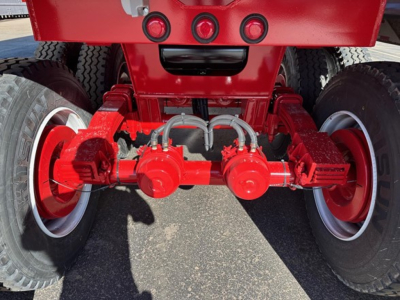 Rear axle assembly of a red dump trailer showing red-painted axles, dual wheels, and braking components with hoses and mounting hardware.