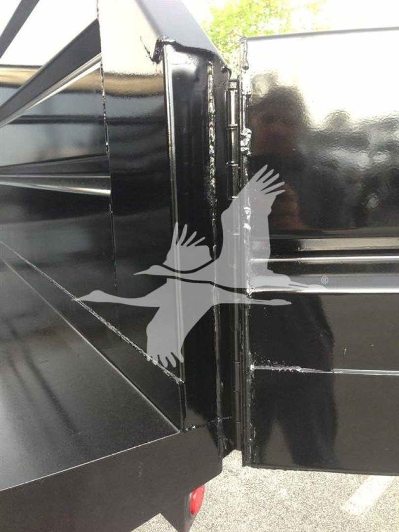 Dump truck trailer hitch area showing black metal structure with welded seams and a logo of a flying bird overlaid. Part of the dump bed and hinge area visible.