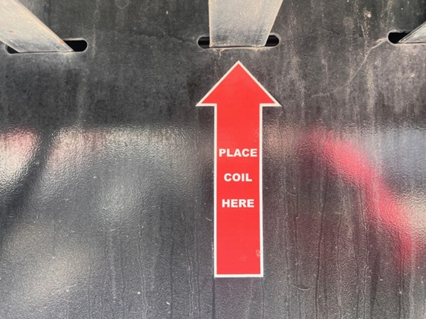 Tall red arrow sticker on trailer reading PLACE COIL HERE on black surface; parts of metal structure visible above.