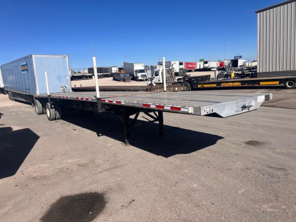 Flatbed trailer with a blue toolbox at the front, long aluminum deck, air suspension legs, and reflective side markings, viewed from side in a yard.