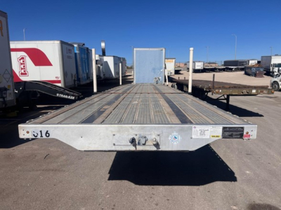 Transcraft flatbed trailer rear view showing metal deck, hitch, and clearance lights with other trailers in background.