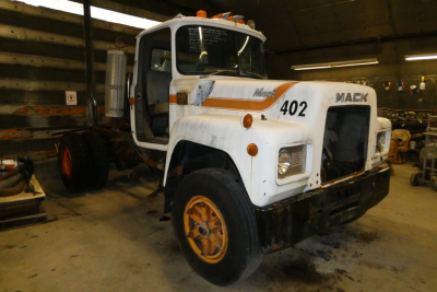 Mack R690T cab and chassis truck in a workshop. Front shows white body with orange and brown stripes, 'Mack' logo on the grille, number '402' on the side, and orange wheels. Other equipment and tires in background.