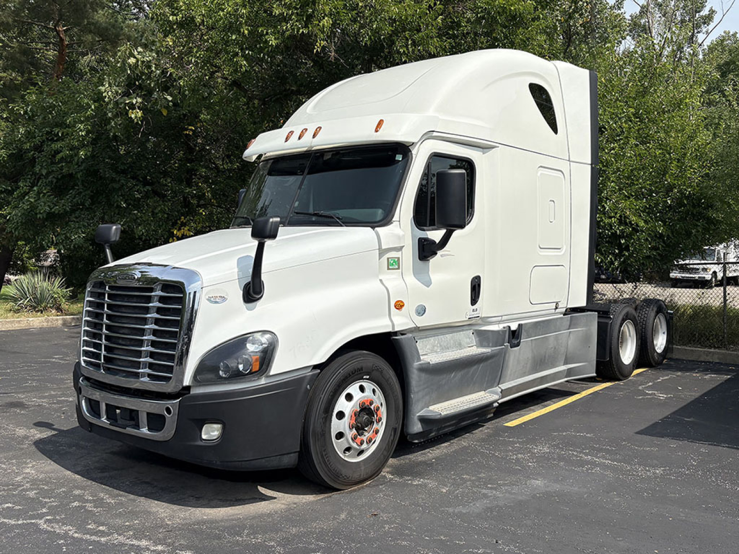 Truck cab: 2018 Freightliner Cascadia125 conventional sleeper, white exterior with aerodynamic raised roof, chrome grille, dual rear axles, driver side mirror, parked on asphalt.