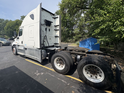 Truck side view showing Freightliner Cascadia125 sleeper with exposed chassis and rear wheels, one cab door, and a parking lot in background.