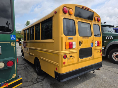 Rear three-quarter view of a yellow school bus with emergency exit sign, red/amber lights, handicap placard, and SLASH text on rear. Visible text includes EMERGENCY DOOR and school bus details plate near tail.