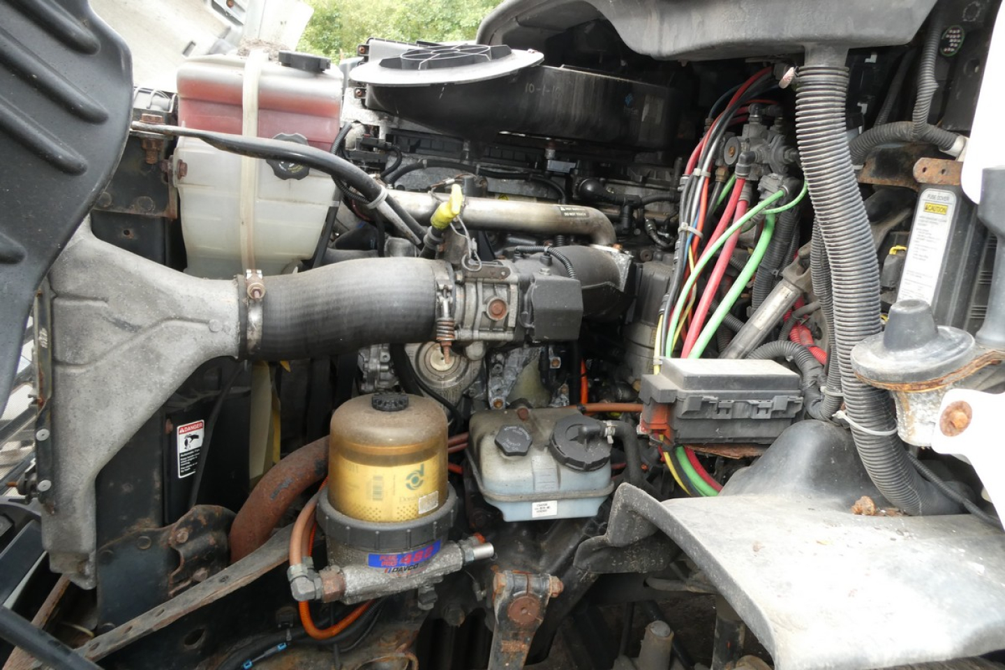 Engine bay of a Freightliner Cascadia 113 daycab showing hoses, wiring harness, coolant reservoir, and various metal components.
