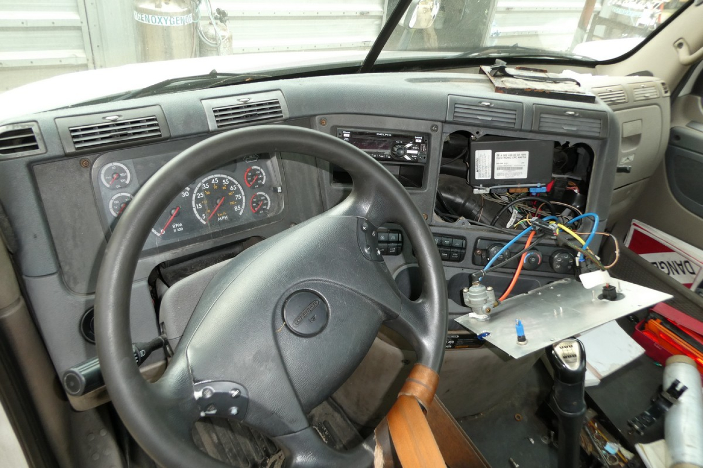 Dashboard and steering wheel of a Freightliner Cascadia cab, with exposed wiring and component panel on the passenger side.