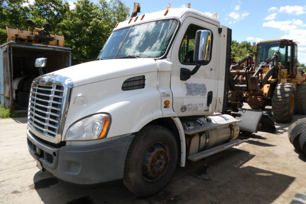 White Freightliner Cascadia 113 daycab truck in a yard, front-left view, missing front wheel, attached heavy equipment behind.