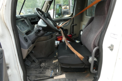 Truck cab interior of a 2012 Freightliner Cascadia 113 showing driver seat, steering wheel, and dash with seat belt laid across seat.