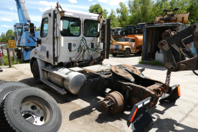 Truck chassis with cab and missing fifth wheel, Freightliner Cascadia 113 rear view showing exposed driveline and bumper, front tire visible, workyard background.