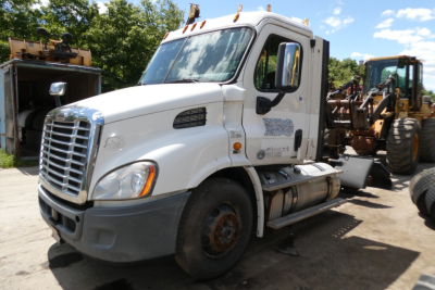 White Freightliner Cascadia 113 daycab truck in a yard, front-left view, missing front wheel, attached heavy equipment behind.