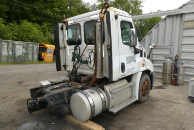 Truck cab of a Freightliner Cascadia 113 daycab tractor with attached fuel tank and rear machinery; side view shows white cab, steps, exhaust, and hitch area.