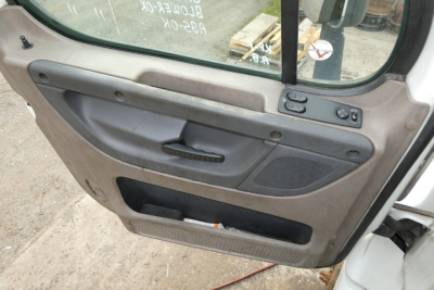 Truck door panel of a 2012 Freightliner Cascadia 113 showing inner door with armrest, window switch, and storage pocket. Visible text: No legible model text; generic interior components.