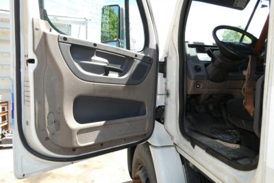 Freightliner Cascadia 113 cab interior and open driver door showing steering wheel and pedals, beige door panel and floor mat.