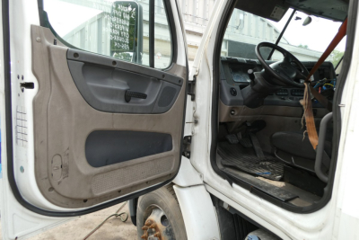 Truck cab interior and door area of a Freightliner Cascadia 113, showing open driver door with beige panel, steering wheel, and dashboard.