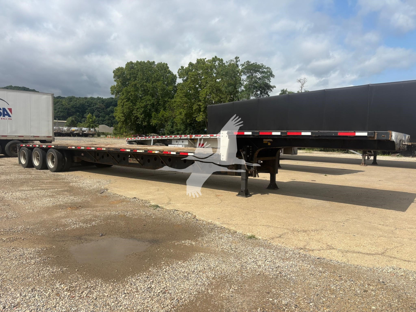 2015 Transcraft DTL-3000 drop deck trailer with extended flatbed and landing gear visible; there is a raised black deck, white support legs, and a partial trailer in background.