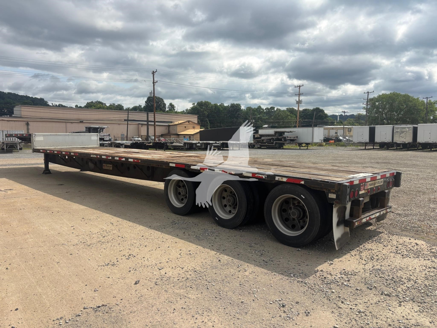 Trailer deck showing a Transcraft DTL-3000 flatbed trailer with dual rear axles and side marker lights; empty bed, gravel lot, overcast sky.