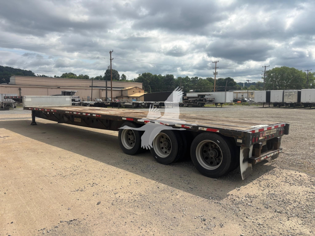 Trailer deck showing a Transcraft DTL-3000 flatbed trailer with dual rear axles and side marker lights; empty bed, gravel lot, overcast sky.