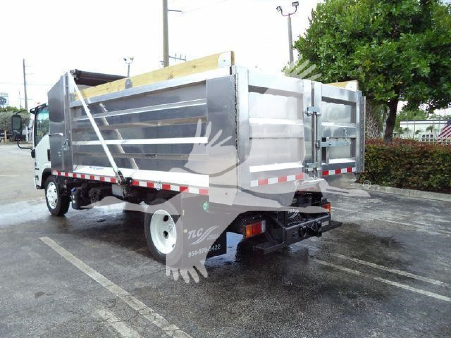 Dump truck rear and side view showing Isuzu NPR-XD dump body with metal sides and tailgate, reflective tape along lower edge, white cab, and rear bumper equipment.