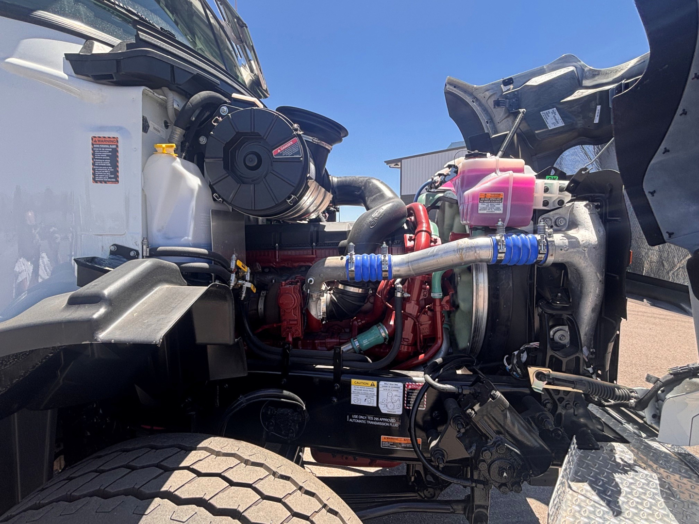 Truck engine bay of a 2026 Mack GR64B showing engine with red components, hoses, coolant reservoir, and turbo assembly.
